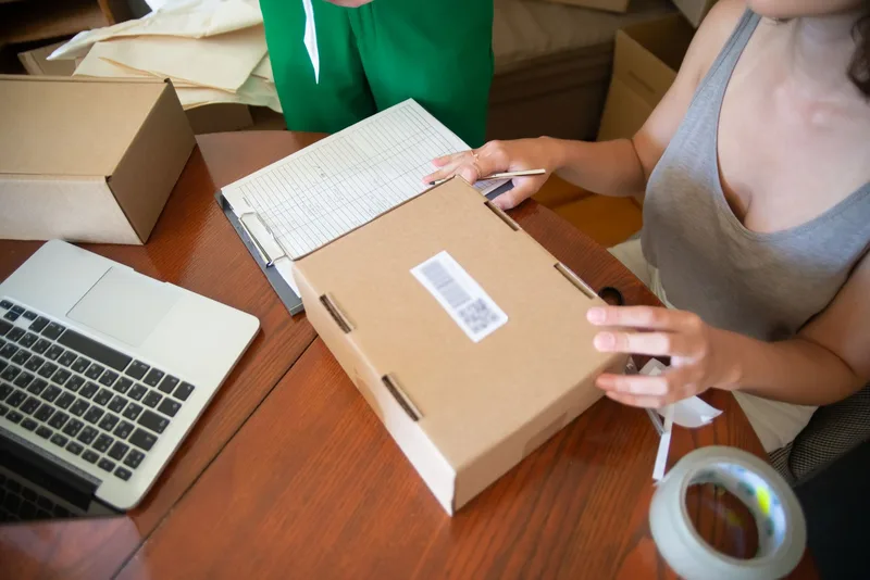 Seller taping a parcel on a wooden desk beside a laptop and order sheet